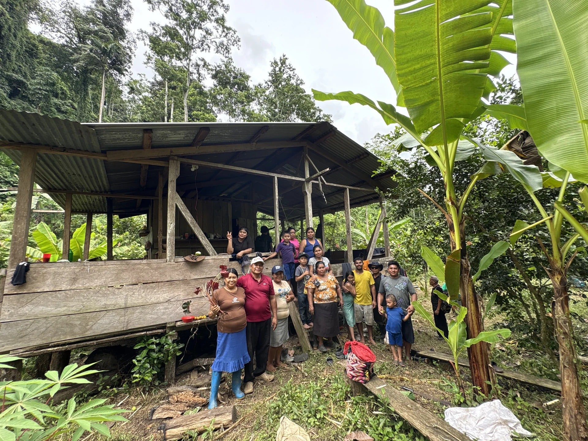 A group of adults and children gathered beside a wooden home among trees.