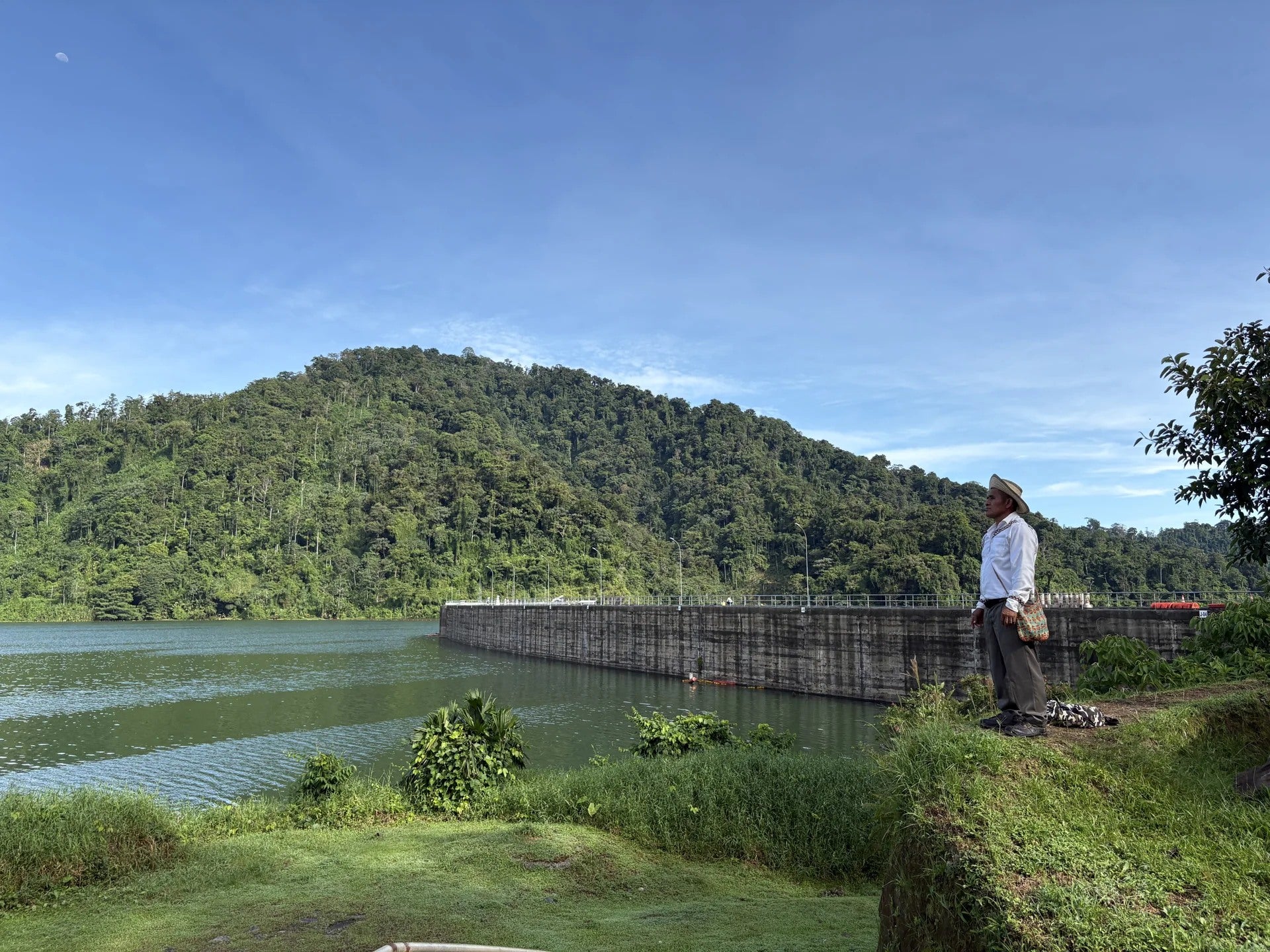 A man stands beside the dam and water, with forested hills behind him.