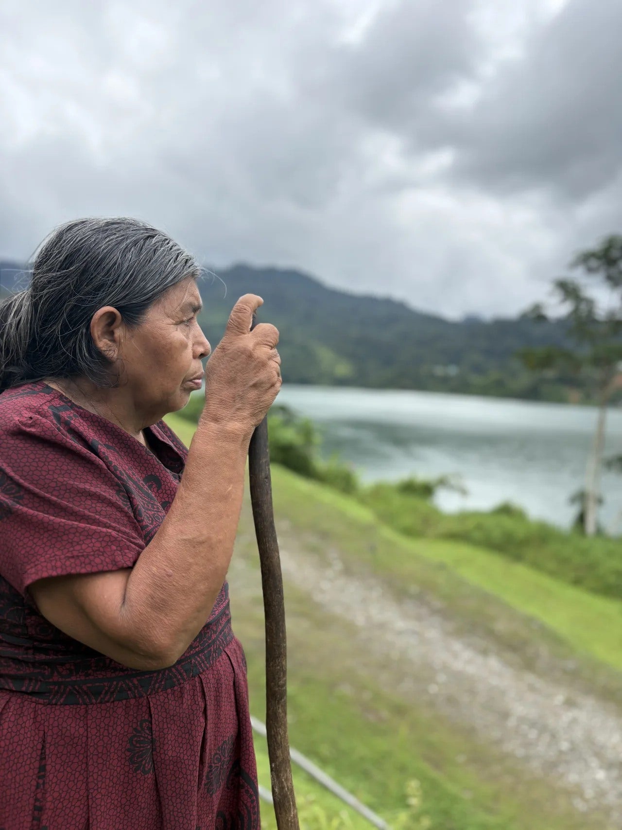 A woman in a burgundy dress holds a wooden staff beside the water.